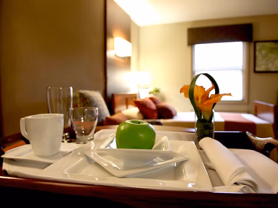 A close-up view of a tray with a green apple on a small white plate, a white coffee cup, two empty glasses, a rolled white napkin, and a small vase with orange flowers. In the background, there is a bed with pillows and a window with a brown valance in a softly lit room.