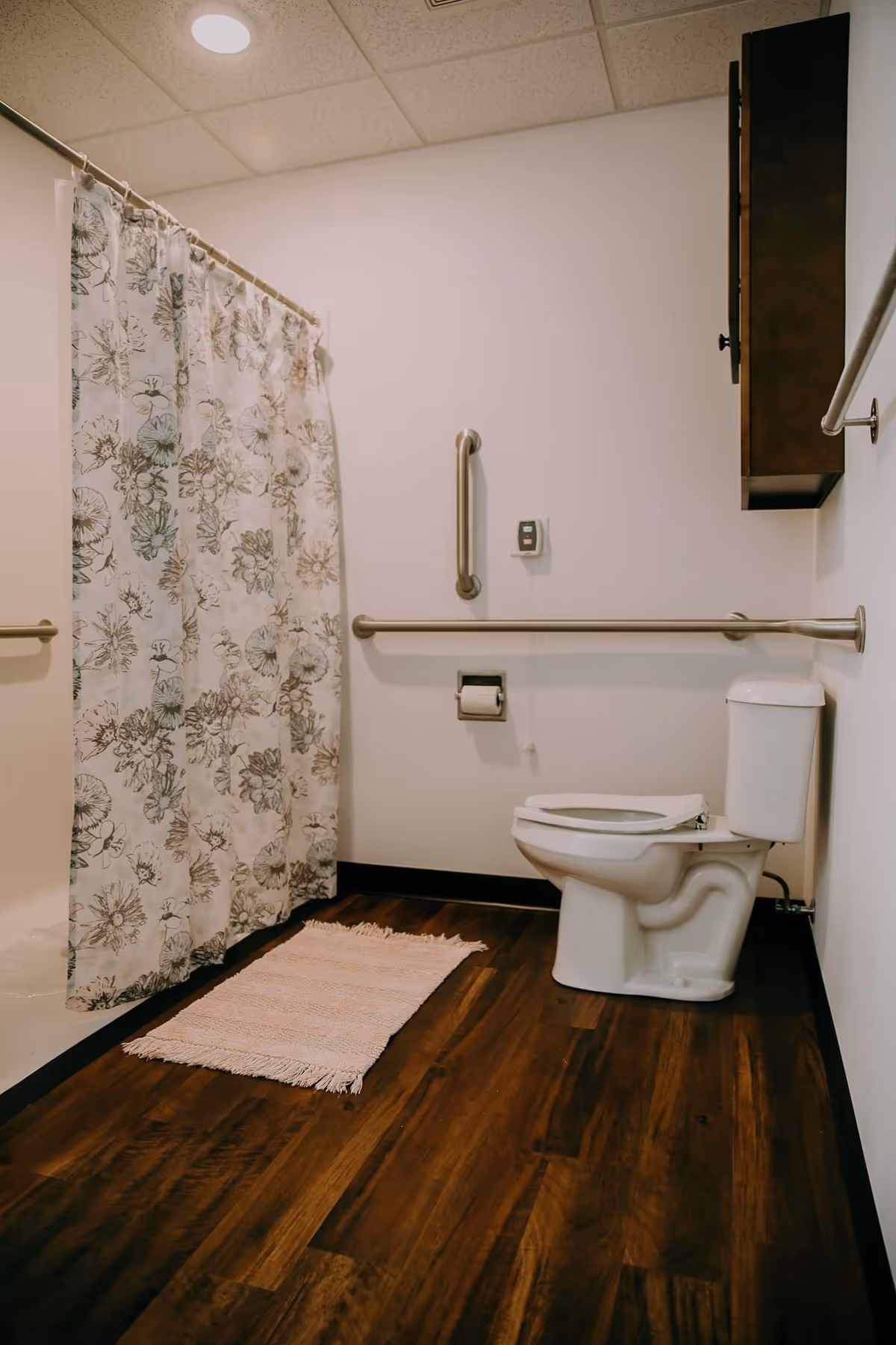 Accessible bathroom with a toilet, wall-mounted grab bars, floral shower curtain, and wood-look flooring.