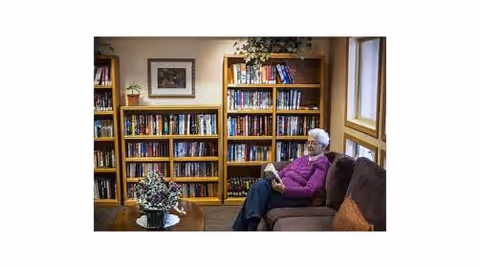An elderly woman wearing a purple sweater sits on a brown couch reading a book in a cozy room with bookshelves filled with books and a framed picture on the wall. A small table with a floral arrangement is in front of her.
