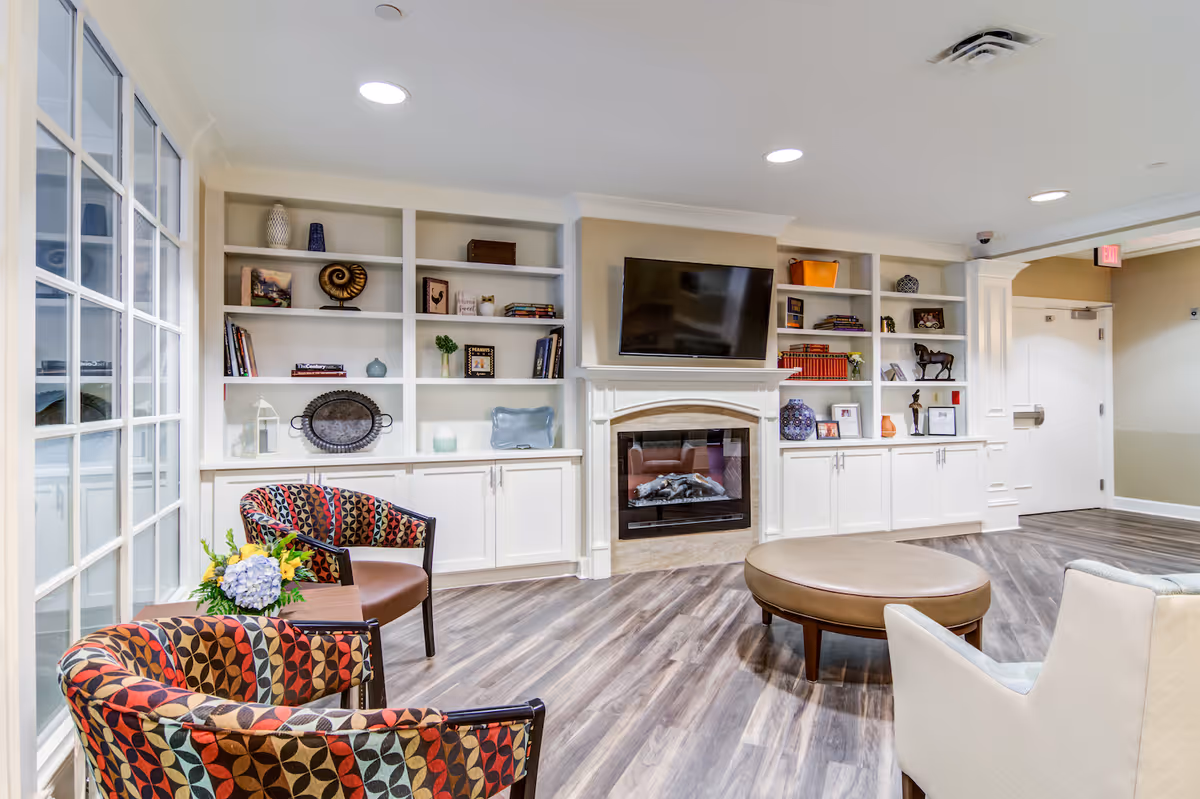 A bright and cozy living room area featuring built-in white shelves filled with decorative items and books, a wall-mounted flat-screen TV above a fireplace, patterned armchairs with a small table holding a flower arrangement, a round leather ottoman, and a white upholstered chair on wood flooring.