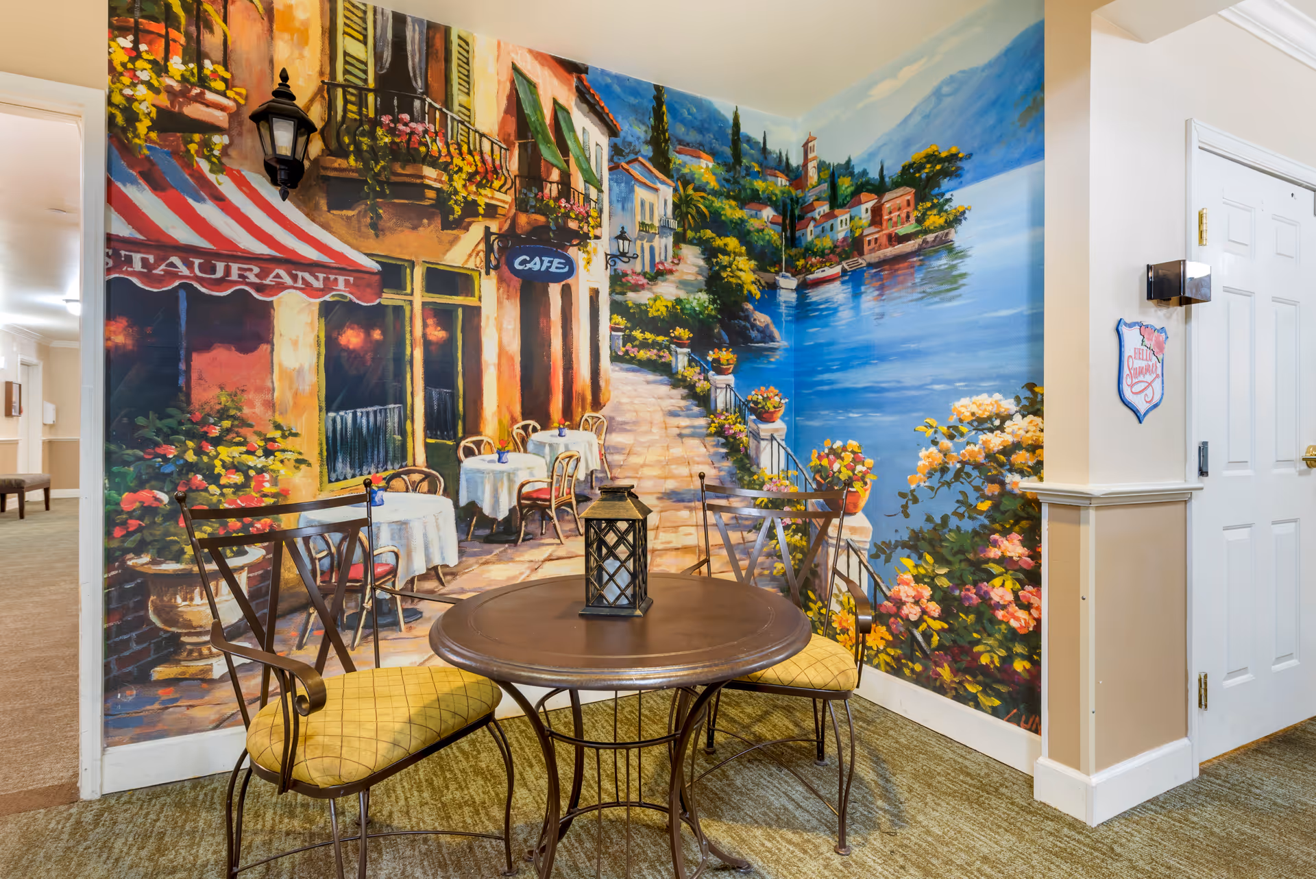A small seating area with a round metal table and two metal chairs with yellow cushions is positioned in front of a colorful mural depicting a scenic outdoor café by the water with flowers and buildings. A lantern sits on the table. The area is carpeted and adjacent to a hallway with beige walls and a white door.