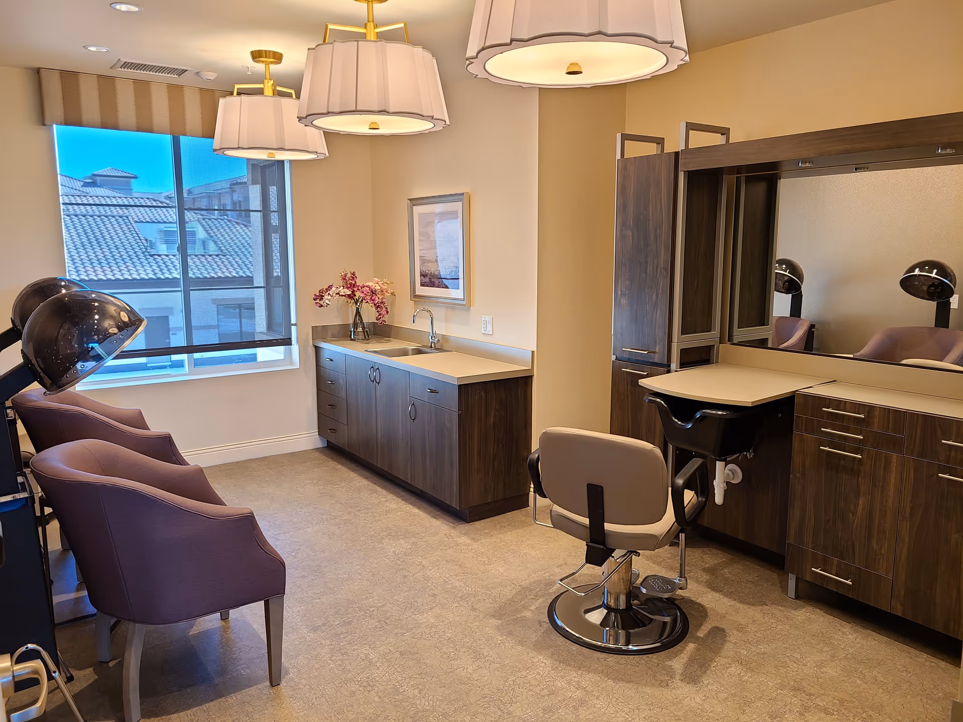Interior view of a salon area in a senior living facility with a large mirror, salon chair, hair drying stations, purple armchairs, a countertop with a sink, and a window showing a building with a tiled roof outside.