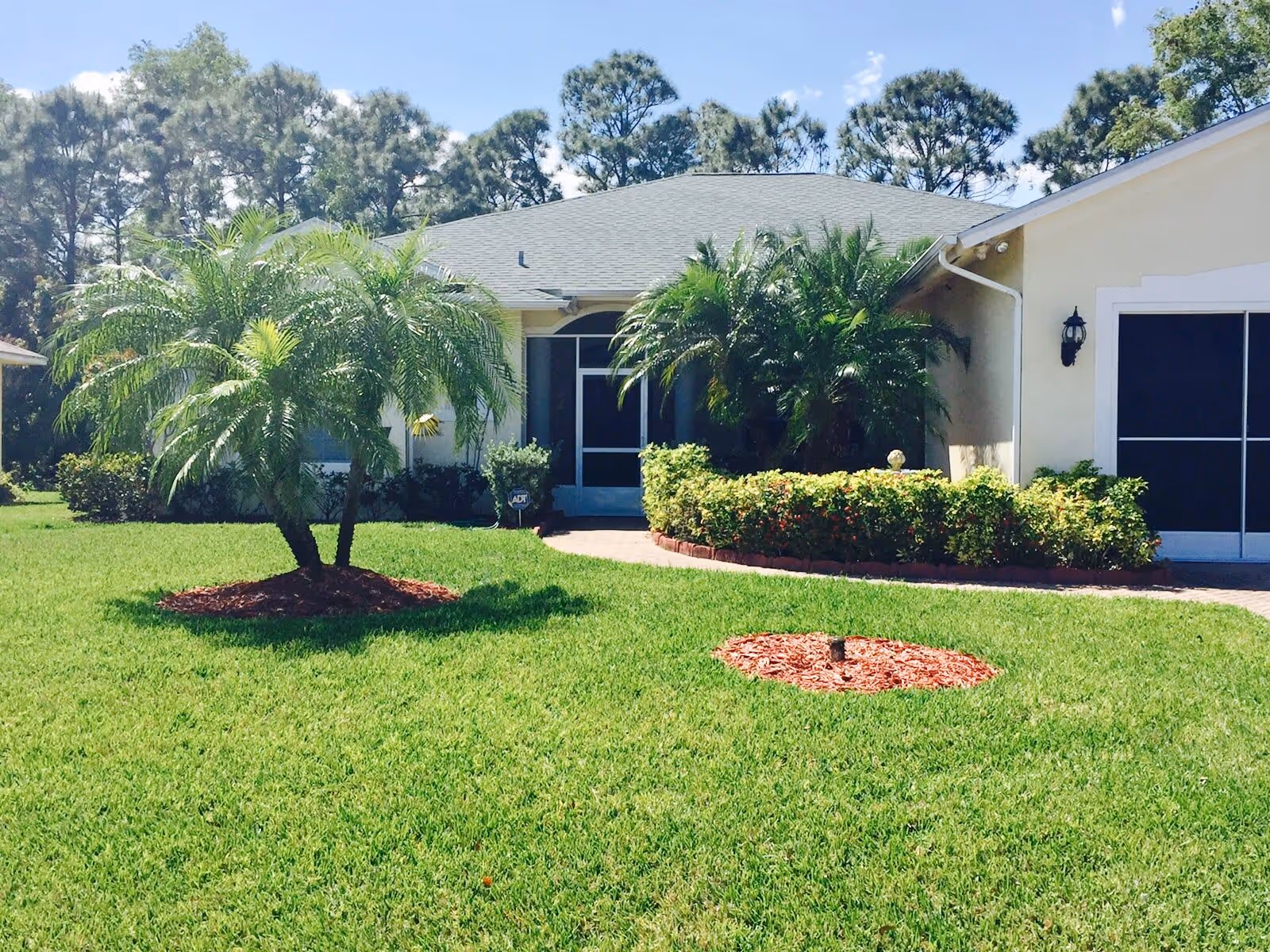Front exterior view of a single-story residential building with a well-maintained green lawn, palm trees, shrubs, and a paved walkway leading to a screened front door under a clear blue sky.