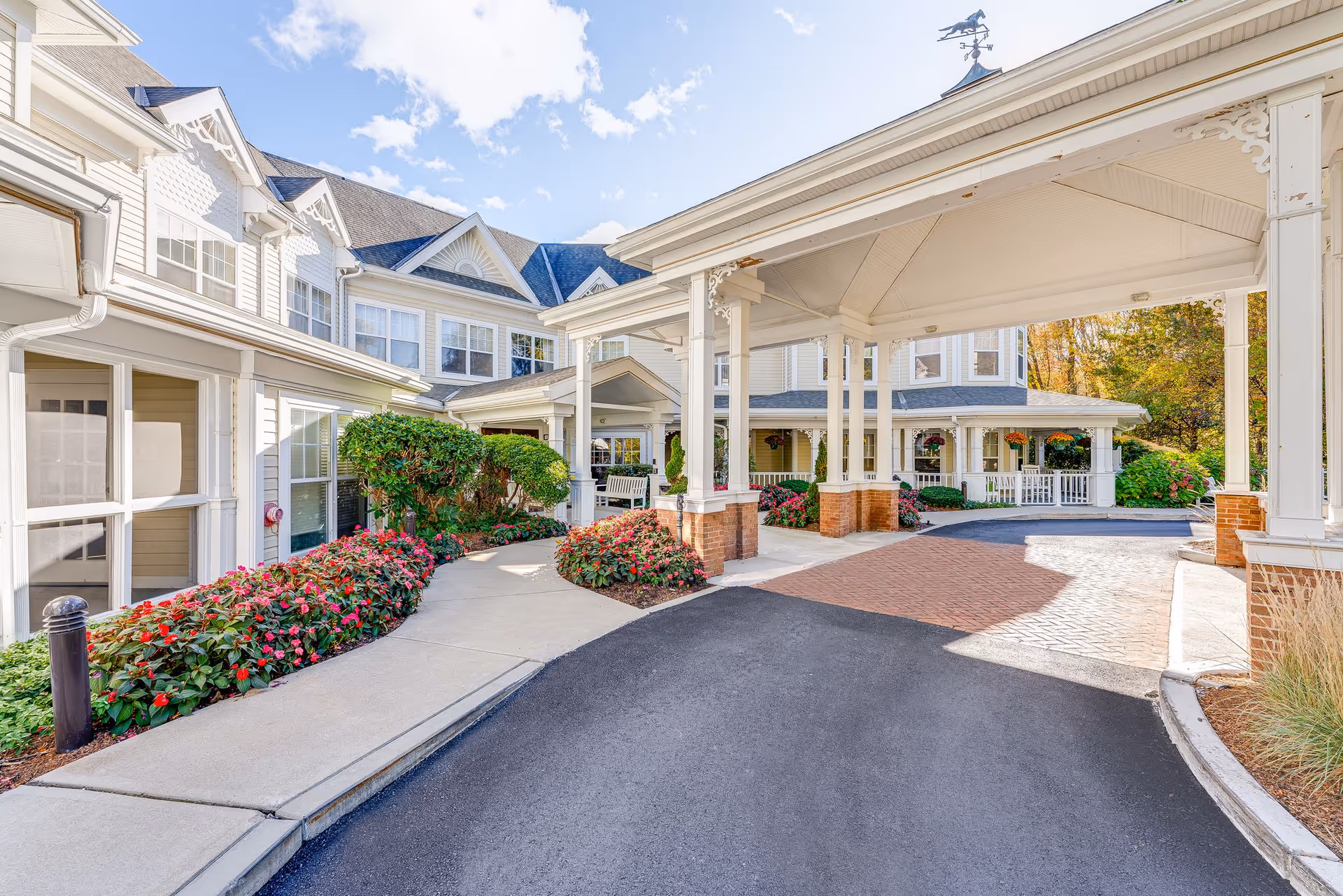 Entrance area of Sunrise of Wayland senior living facility with a covered drop-off zone, paved driveway, flower beds with red flowers, bushes, and a white multi-story building with large windows under a partly cloudy sky.