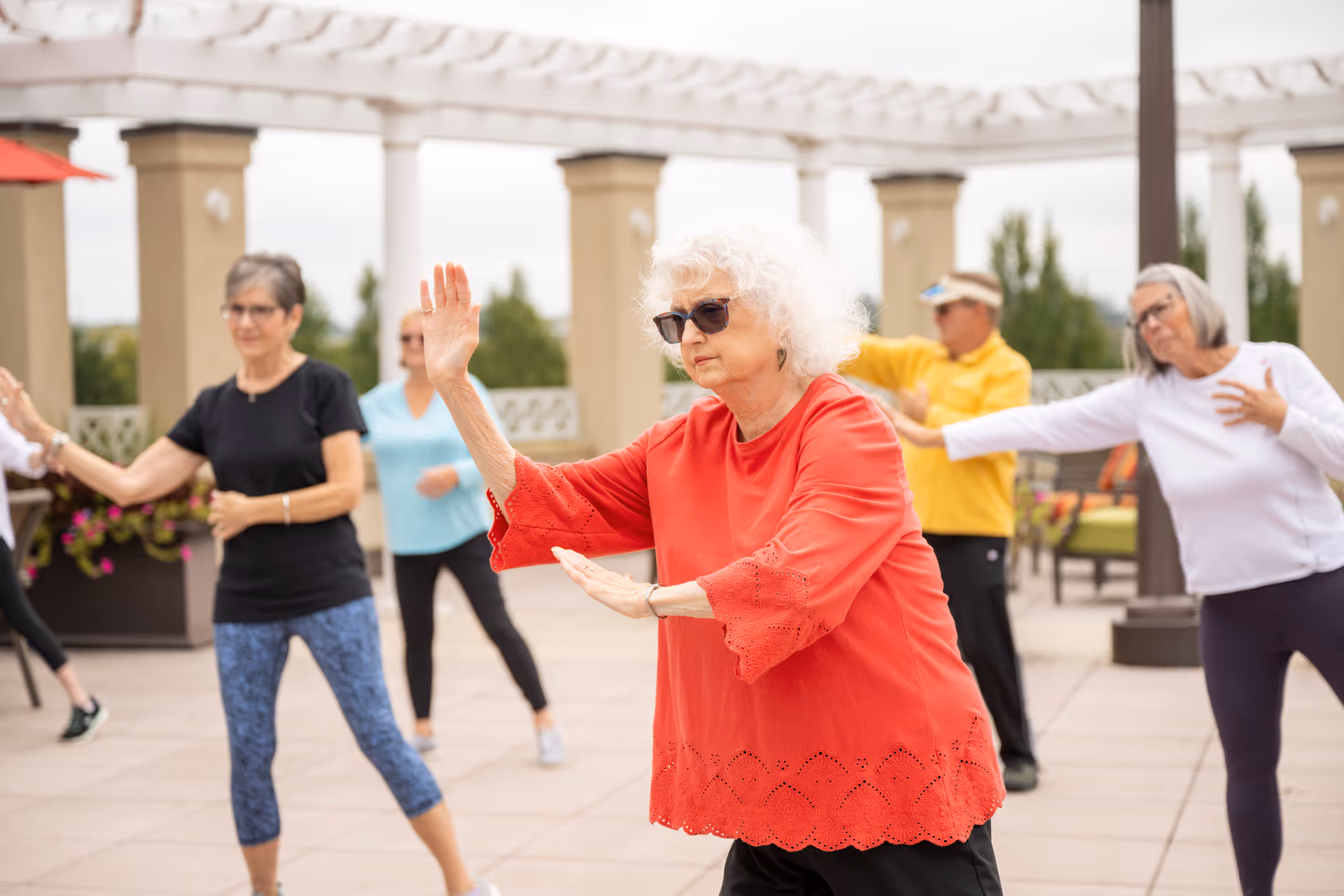 A group of elderly people practicing Tai Chi outdoors on a patio with columns and greenery in the background. The woman in the foreground is wearing a red top and sunglasses, and others are dressed casually in activewear.