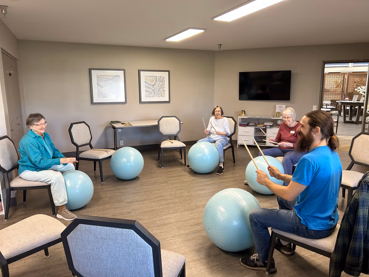 A group of older adults and an instructor sit in a circle in a common room using large exercise balls and drumsticks for a seated group activity.