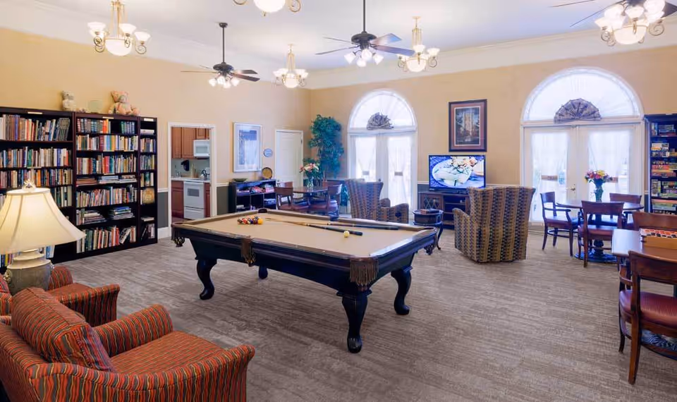 A spacious common area with a pool table in the center, surrounded by comfortable armchairs and tables with chairs. There are bookshelves filled with books on the left side, a television on a stand near large arched windows with sheer curtains, and ceiling fans with lights overhead. The room has a warm, inviting atmosphere with beige walls and carpeted floors.