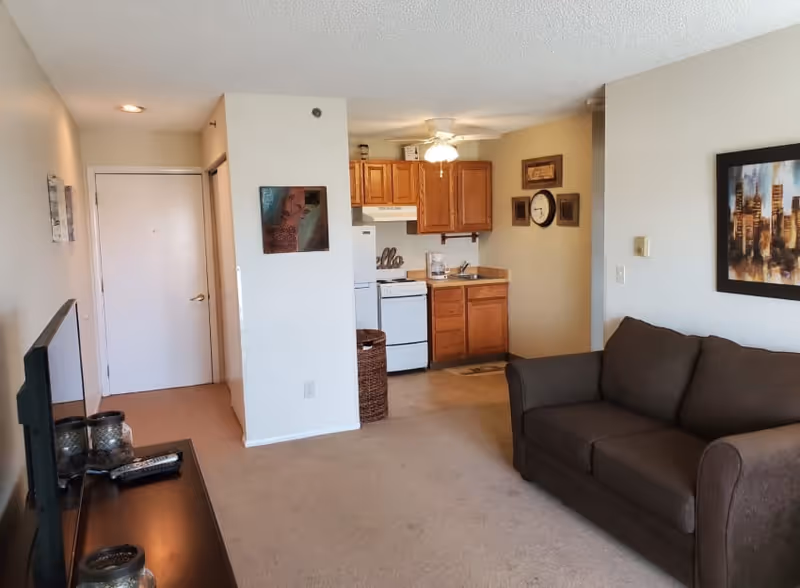 Interior view of a senior living facility apartment showing a small kitchen area with wooden cabinets, a white stove, refrigerator, and a coffee maker. Adjacent to the kitchen is a living room with a dark brown sofa and a TV on a stand. The walls are decorated with framed artwork and a clock. The floor is carpeted and the space is well-lit with ceiling lights.