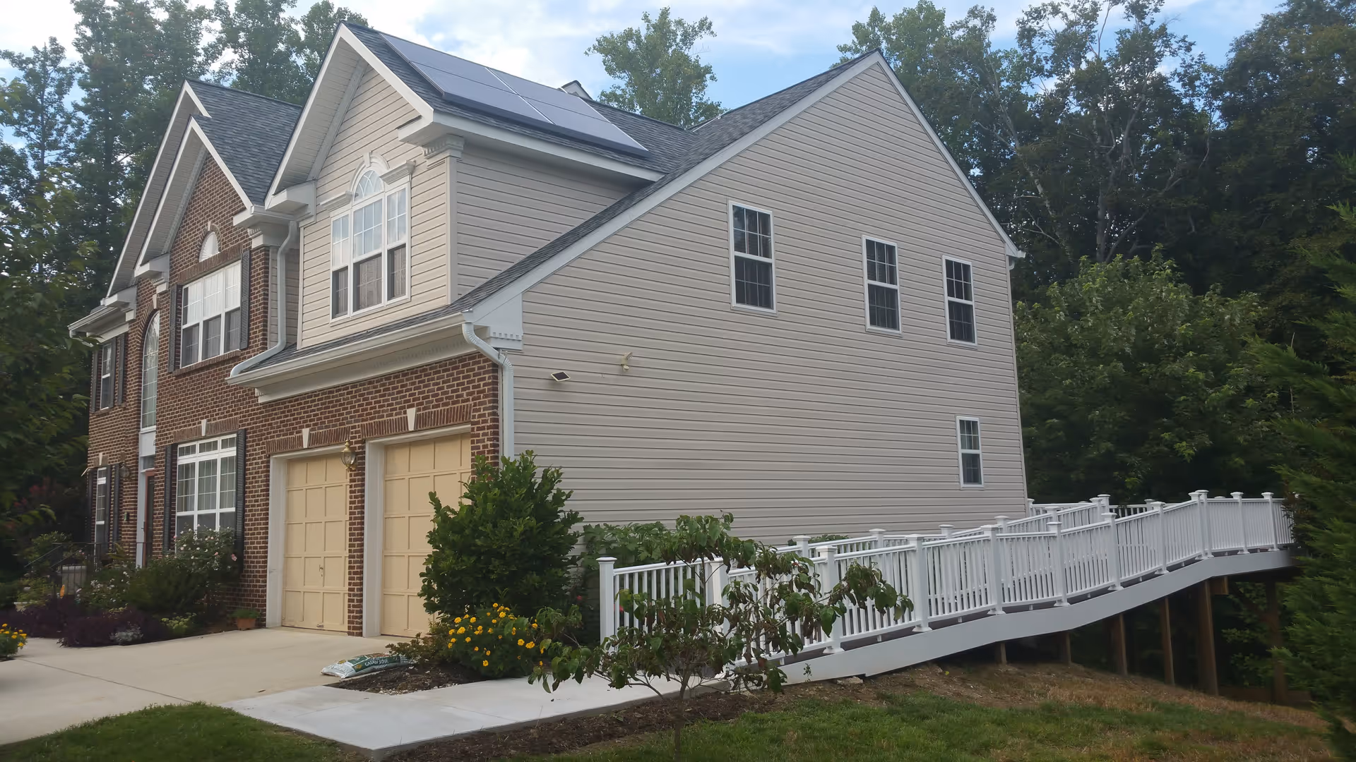 Exterior view of a two-story residential building with a brick and beige siding facade, multiple windows, a double garage, and a white wheelchair-accessible ramp leading to the entrance. The building is surrounded by greenery and trees.