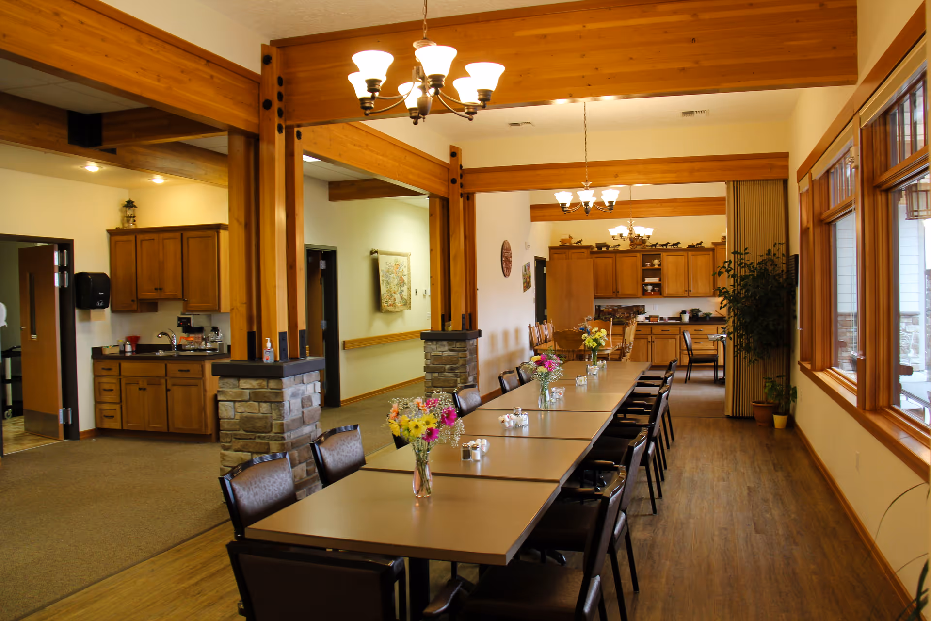 A long dining table with chairs on both sides, decorated with vases of colorful flowers, situated in a spacious room with wooden beams and large windows letting in natural light. The room includes a kitchen area with wooden cabinets and countertops in the background.