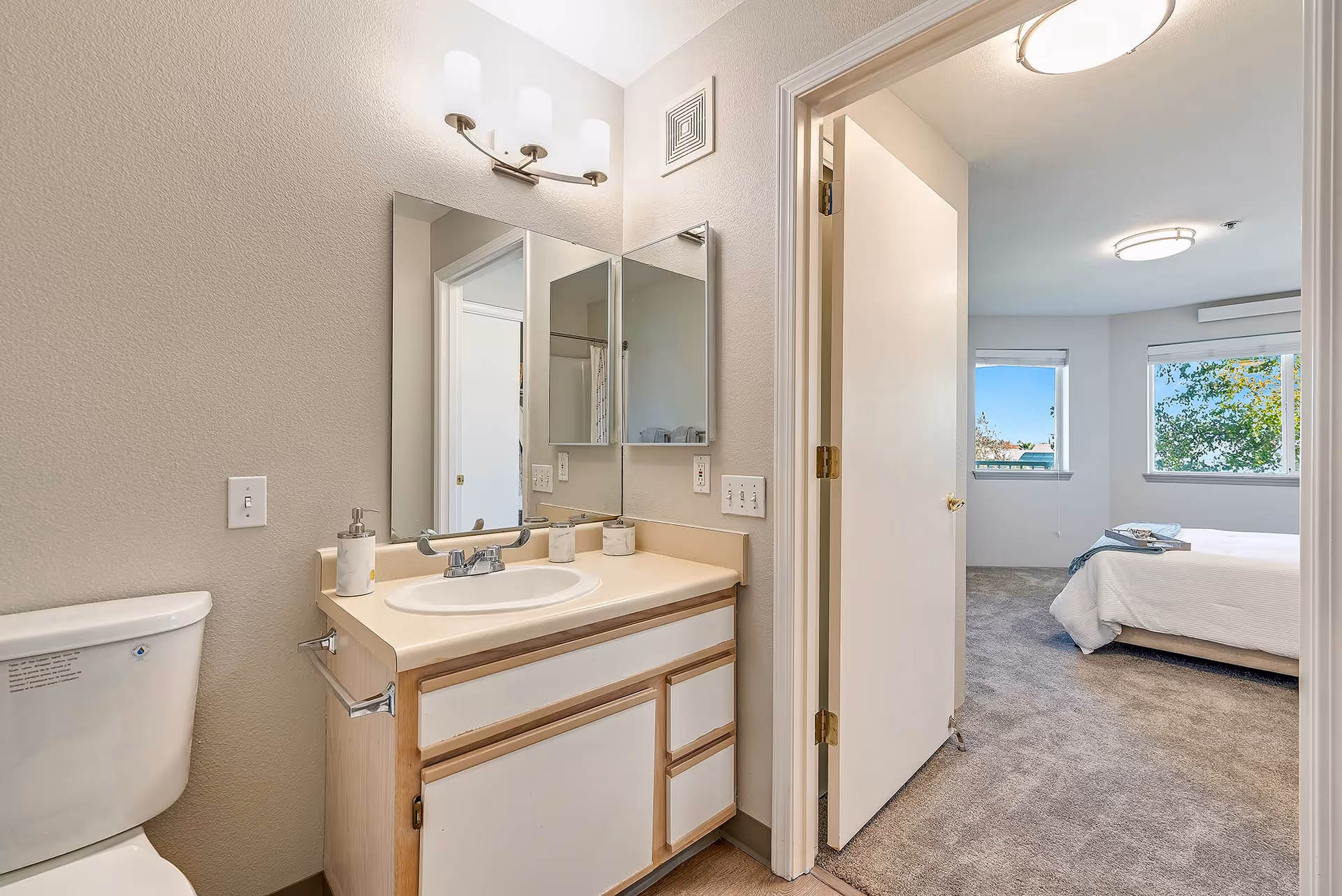 Bathroom with a vanity, sink, mirror and toilet opening into a bedroom with a bed and windows.