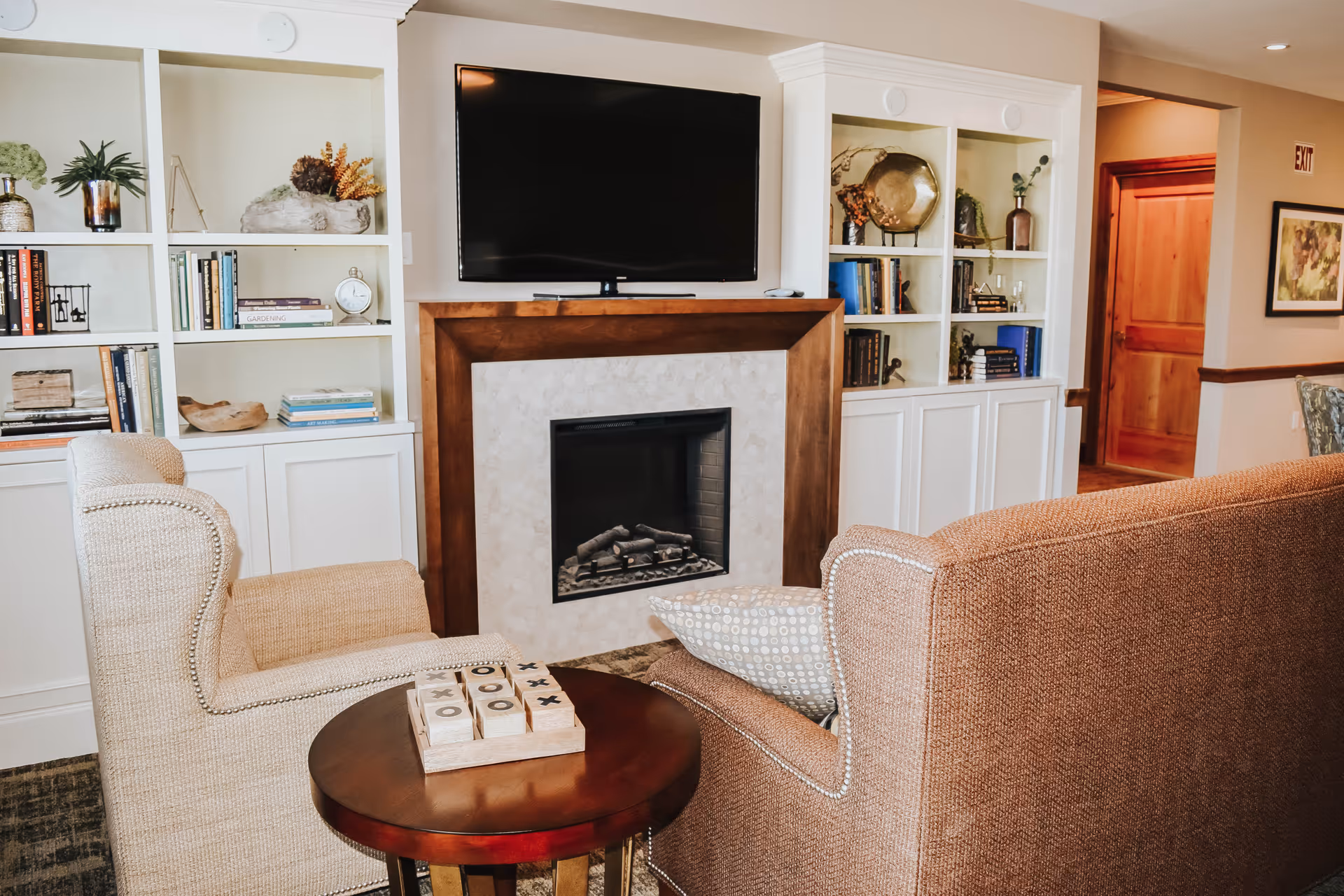 Cozy communal living room with upholstered armchairs around a fireplace, built-in bookshelves, a round side table, and a wall-mounted TV.