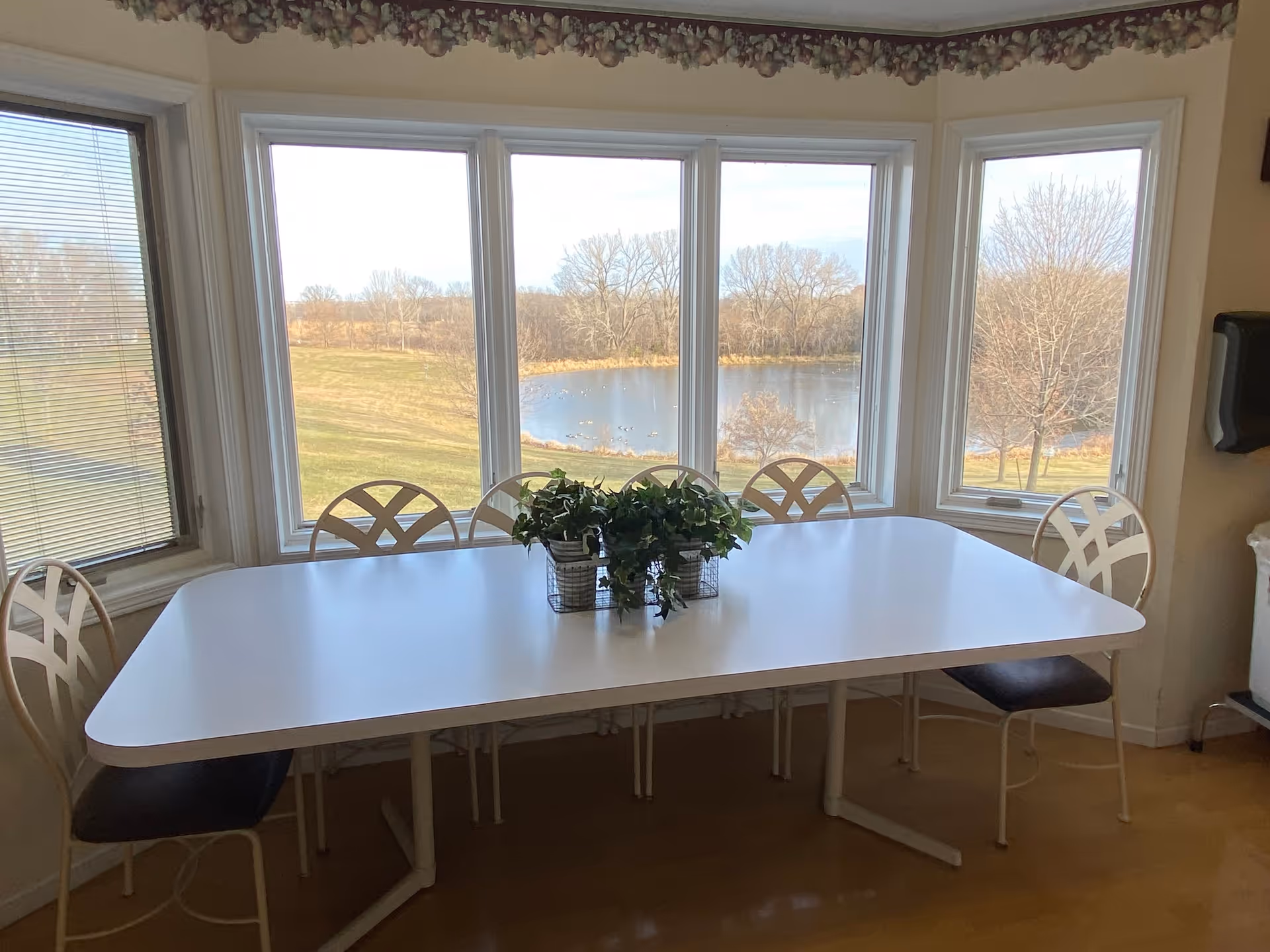 A white rectangular table with five chairs around it is positioned in front of large windows showing a scenic view of a pond and leafless trees outside. There are three small potted plants in wire baskets placed on the table. The room has light-colored walls and a floral border near the ceiling.