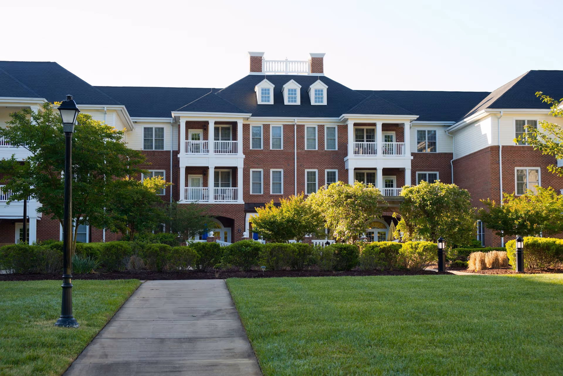 Exterior view of a multi-story senior living facility building with red brick and white siding, featuring balconies, windows, and a well-maintained lawn with bushes and trees in front. A paved walkway leads up to the building, flanked by lamp posts.