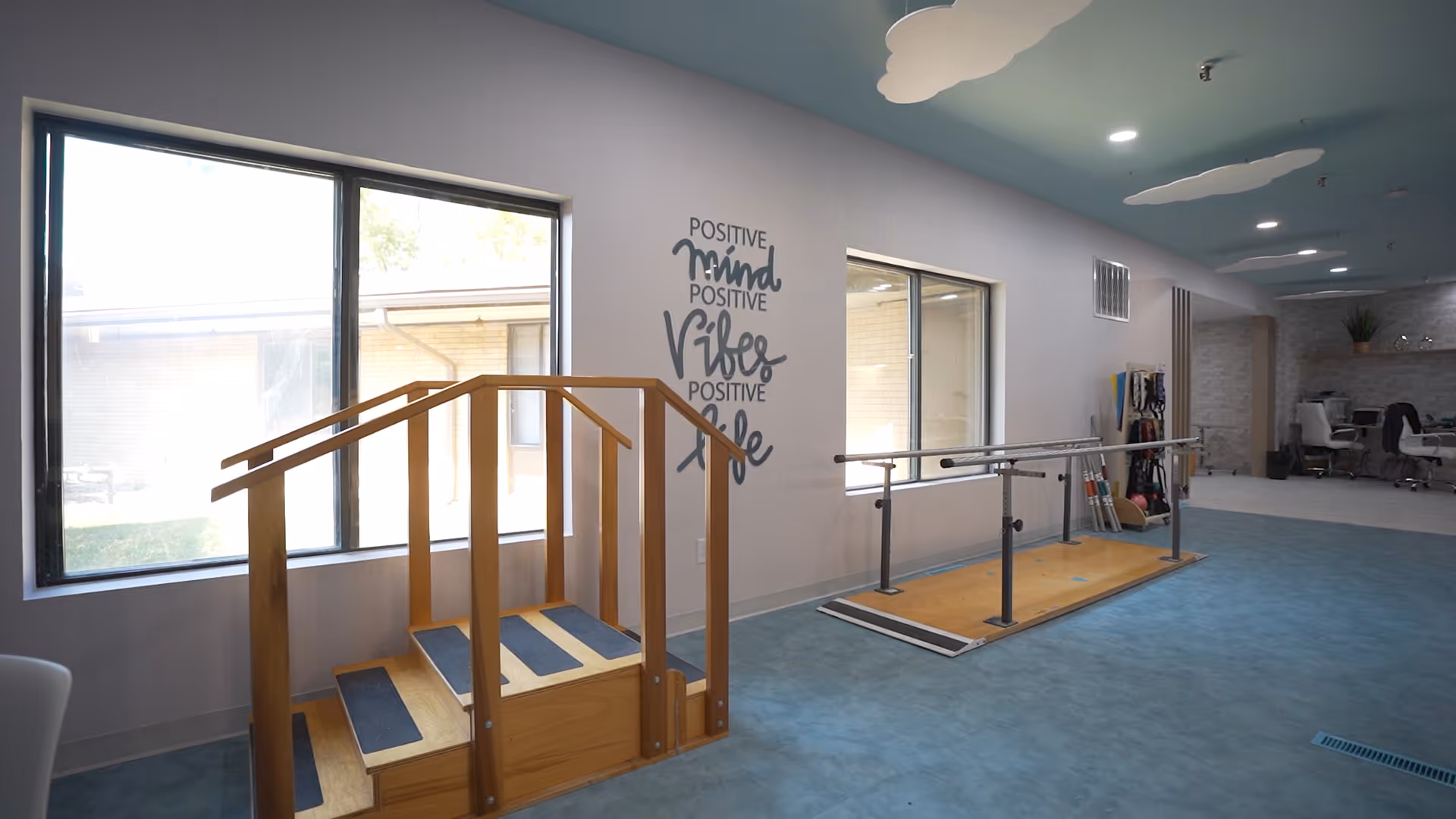 Interior view of a rehabilitation or physical therapy room with wooden stairs and parallel bars for walking exercises. The room has large windows letting in natural light, a blue floor, and a wall decal that reads 'Positive mind, Positive vibes, Positive life.' In the background, there are chairs and therapy equipment.