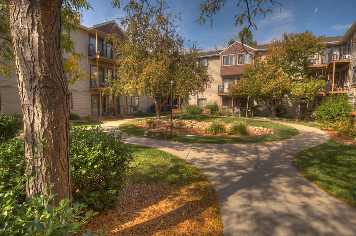 Outdoor courtyard area of a senior living facility with a circular walking path, benches, trees, and shrubs surrounded by a three-story building with balconies and windows under a clear blue sky.