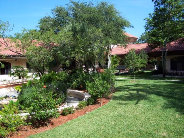 A landscaped outdoor garden area with green grass, various trees, shrubs, and plants. The garden is surrounded by buildings with red-tiled roofs and beige walls under a clear blue sky.