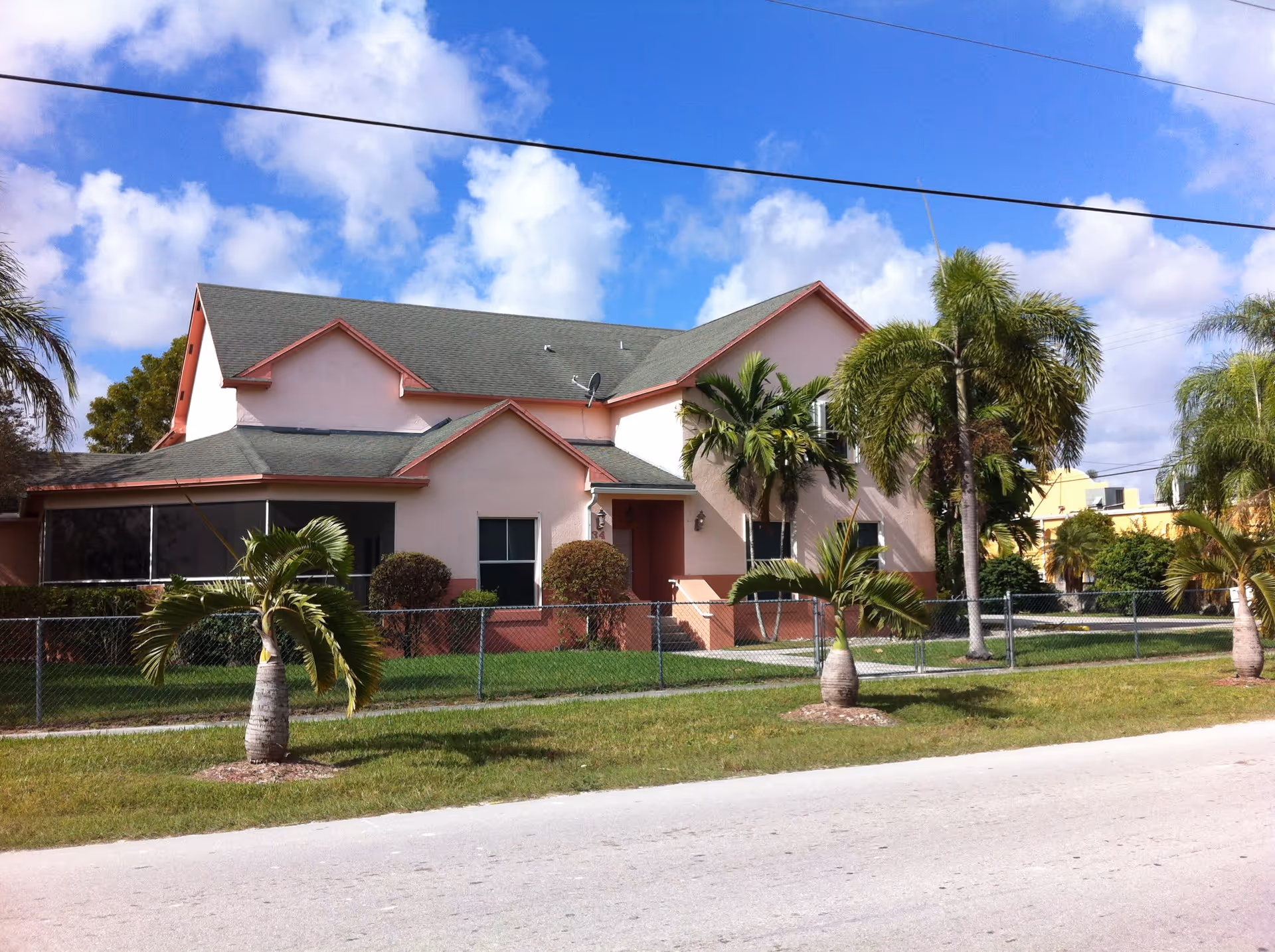Exterior view of a two-story residential building with a green roof and light pink walls, surrounded by a chain-link fence and palm trees under a partly cloudy blue sky.
