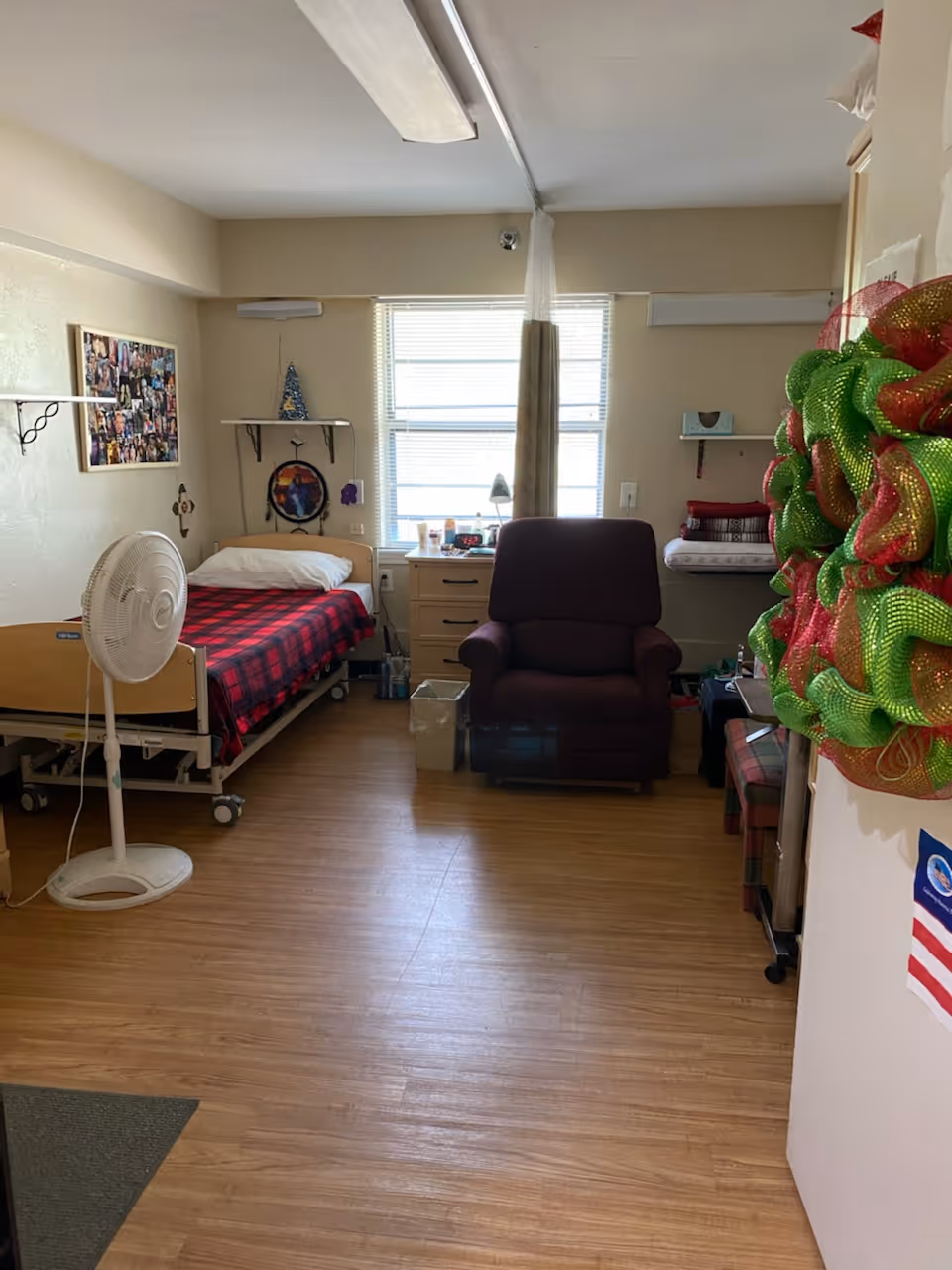 A nursing center room with a single bed covered in a red and black checkered blanket, a white pillow, and a standing fan next to it. There is a dark purple recliner chair in front of a window with blinds and a curtain. A small dresser with various items on top is beside the chair. The room has light-colored walls with shelves holding decorations and folded blankets. A colorful wreath is hanging on the right side near the entrance.