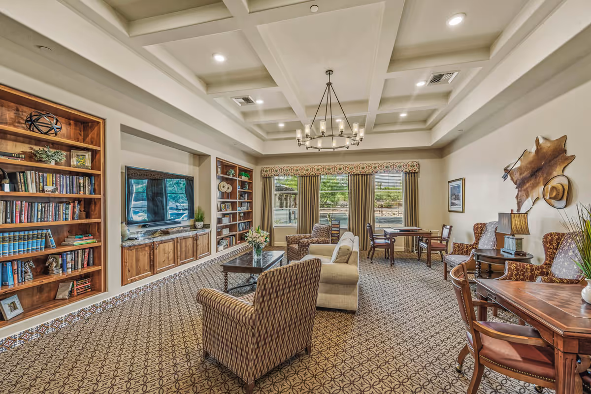 A spacious and well-lit living room with patterned carpet, a beige sofa, two patterned armchairs, wooden bookshelves filled with books and decorative items, a flat-screen TV mounted on the wall, and several wooden tables with chairs. Large windows with curtains allow natural light to enter the room. The ceiling features a coffered design with recessed lighting and a chandelier.