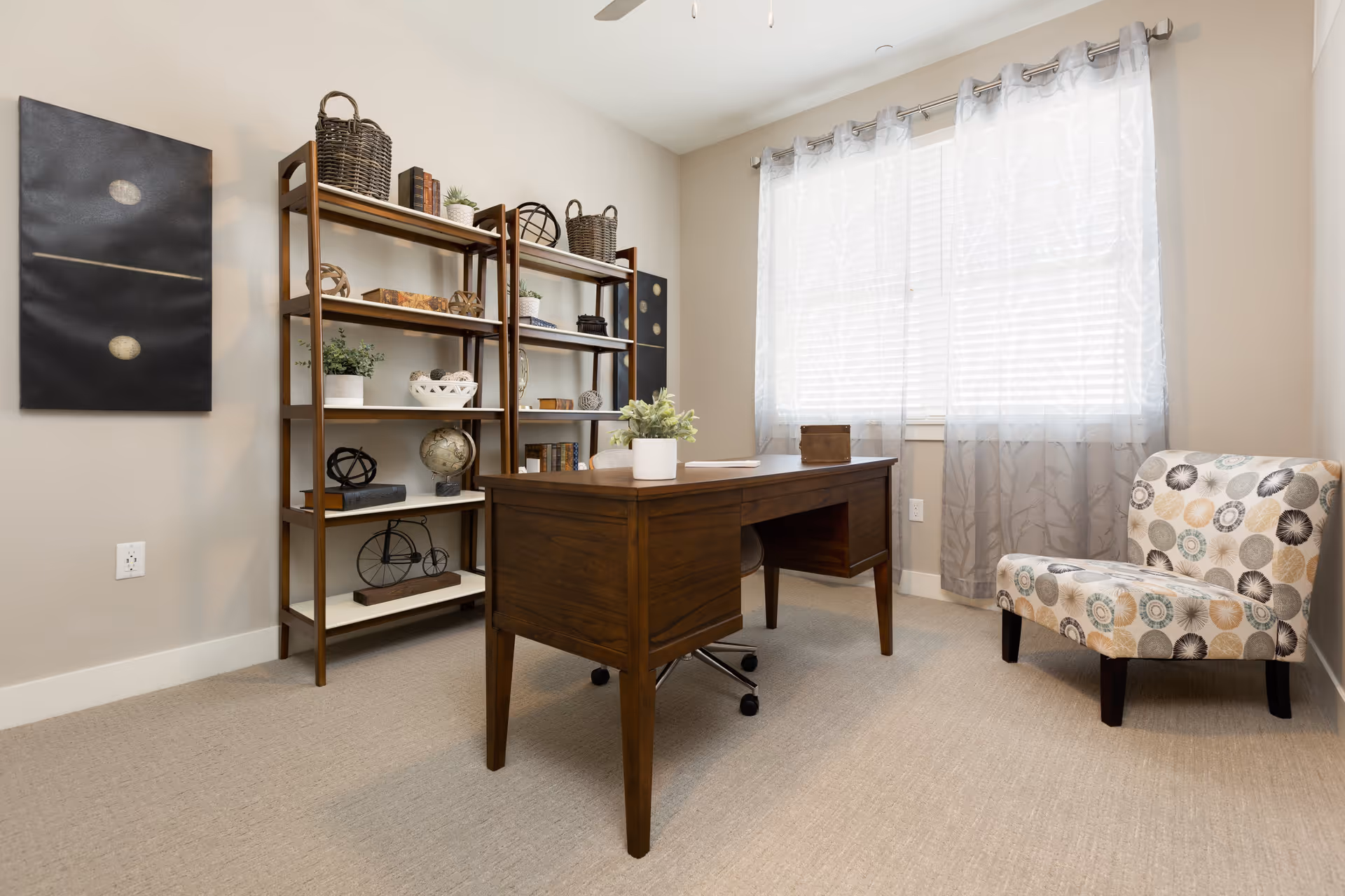A bright office room with a wooden desk and a rolling chair in the center. Behind the desk is a large window with sheer white curtains allowing natural light to fill the room. To the left, there is a tall wooden bookshelf with various decorative items including baskets, books, plants, and globes. On the right side, there is a patterned upholstered chair with circular designs in muted colors. The walls are painted light beige and the floor is carpeted in a neutral tone.