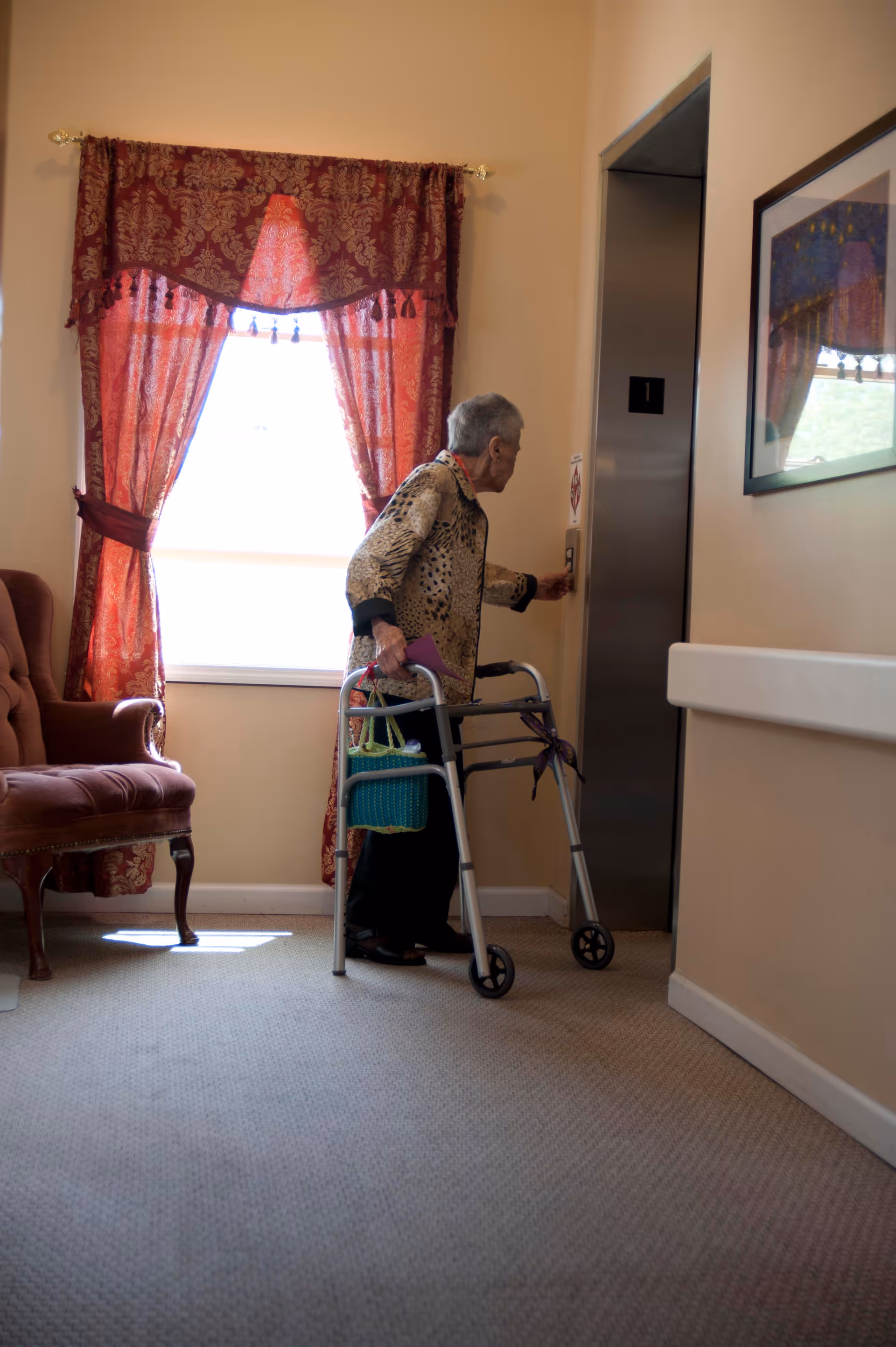 An elderly woman using a walker is pressing the button for an elevator in a hallway. The hallway has beige walls, a window with red patterned curtains, a framed picture on the wall, and a cushioned chair nearby.