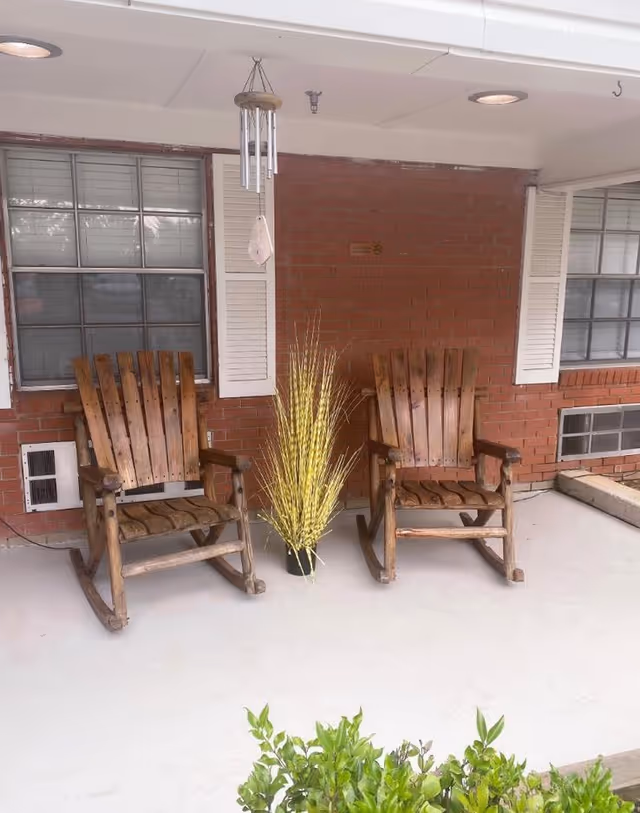 Two wooden rocking chairs on a covered porch with a potted plant between them, a wind chime hanging above, and brick walls with windows in the background.