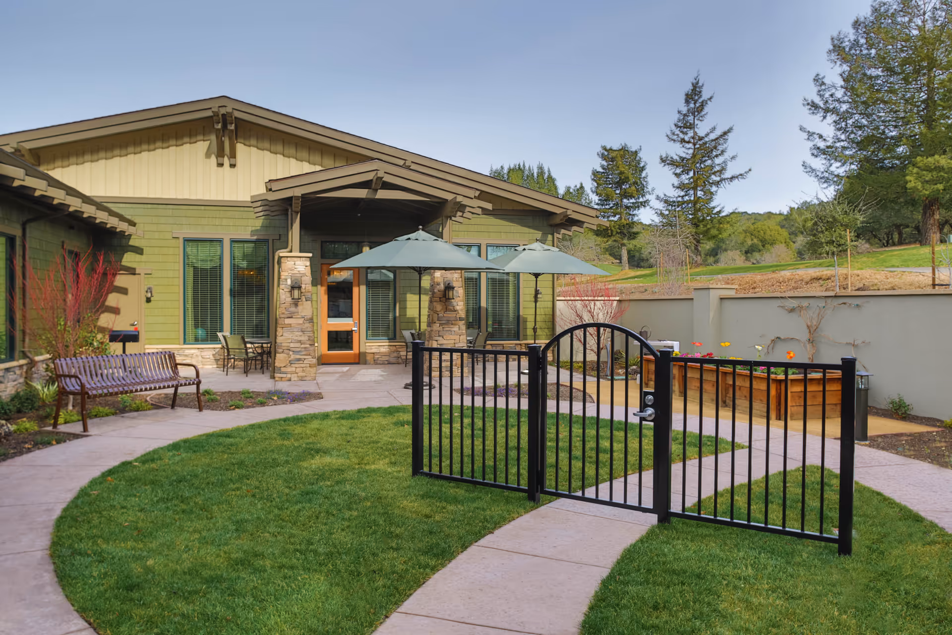 Outdoor courtyard area at The Terraces at Fountaingrove Lodge featuring a green lawn, paved walkways, a black metal gate, wooden bench, patio tables with umbrellas, and a raised garden bed with flowers. The building exterior is green with stone pillars and large windows, surrounded by trees and a clear sky.