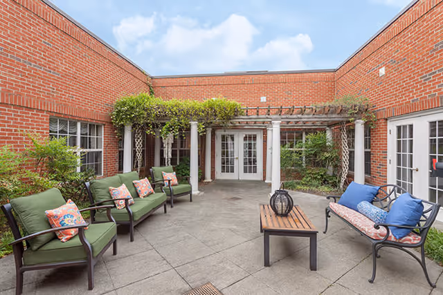 Outdoor patio area with brick walls, green cushioned chairs with colorful pillows, a wooden coffee table, and a metal bench with blue and patterned cushions under a pergola with greenery.