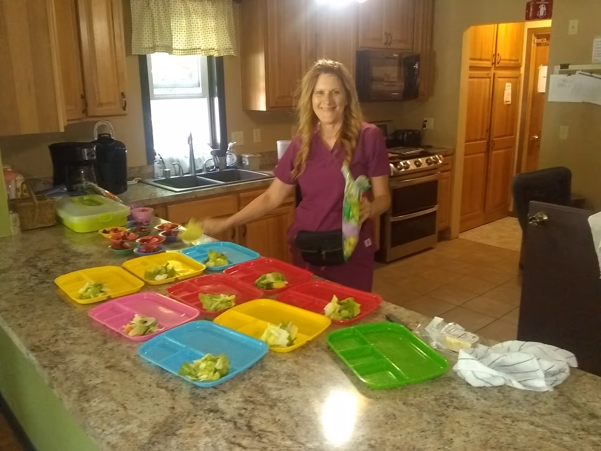 A woman in purple scrubs stands in a kitchen preparing colorful plastic trays with small portions of salad. The kitchen has wooden cabinets, a double sink, a stove, and a microwave. Various food items and kitchen utensils are visible on the countertop.