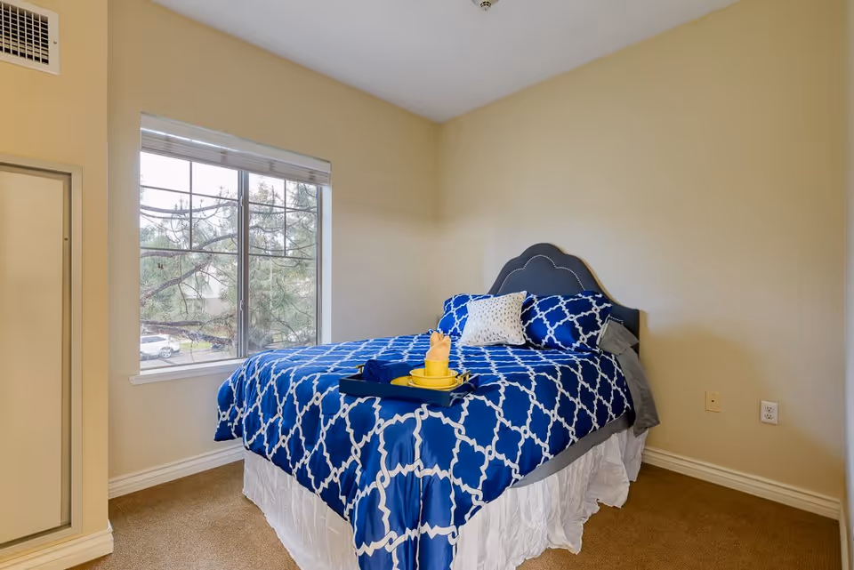 A bedroom with a bed featuring a blue and white patterned comforter and pillows. There is a tray with a yellow cup and rolled towel on the bed. A large window with white blinds shows trees and parked cars outside. The walls are painted beige and the carpet is light brown.