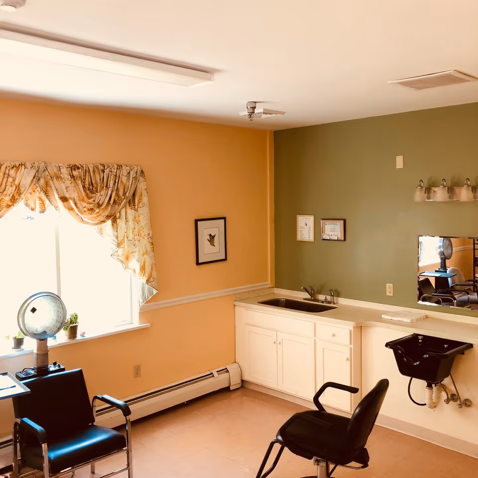 Bright interior salon-style room with two styling chairs, a countertop sink, a black shampoo basin and a window with floral curtains.