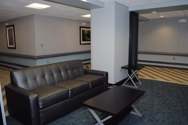 Interior view of a common area in a nursing and rehabilitation center featuring a black leather sofa, a black coffee table, and a small black side table. The room has patterned wallpaper, framed artwork on the walls, and a mix of blue carpet and striped flooring.