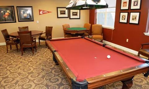 A recreational room with a red pool table in the foreground, a poker table with chairs in the background, framed pictures and a pennant on the walls, and a window letting in natural light.