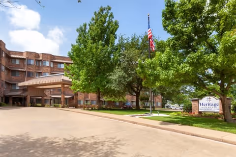 Exterior view of Heritage Place Independent Living facility showing a multi-story brick building with a covered entrance, surrounded by green trees and a lawn. An American flag is flying on a flagpole near the entrance, and a sign with the facility name is visible on the right side of the image.