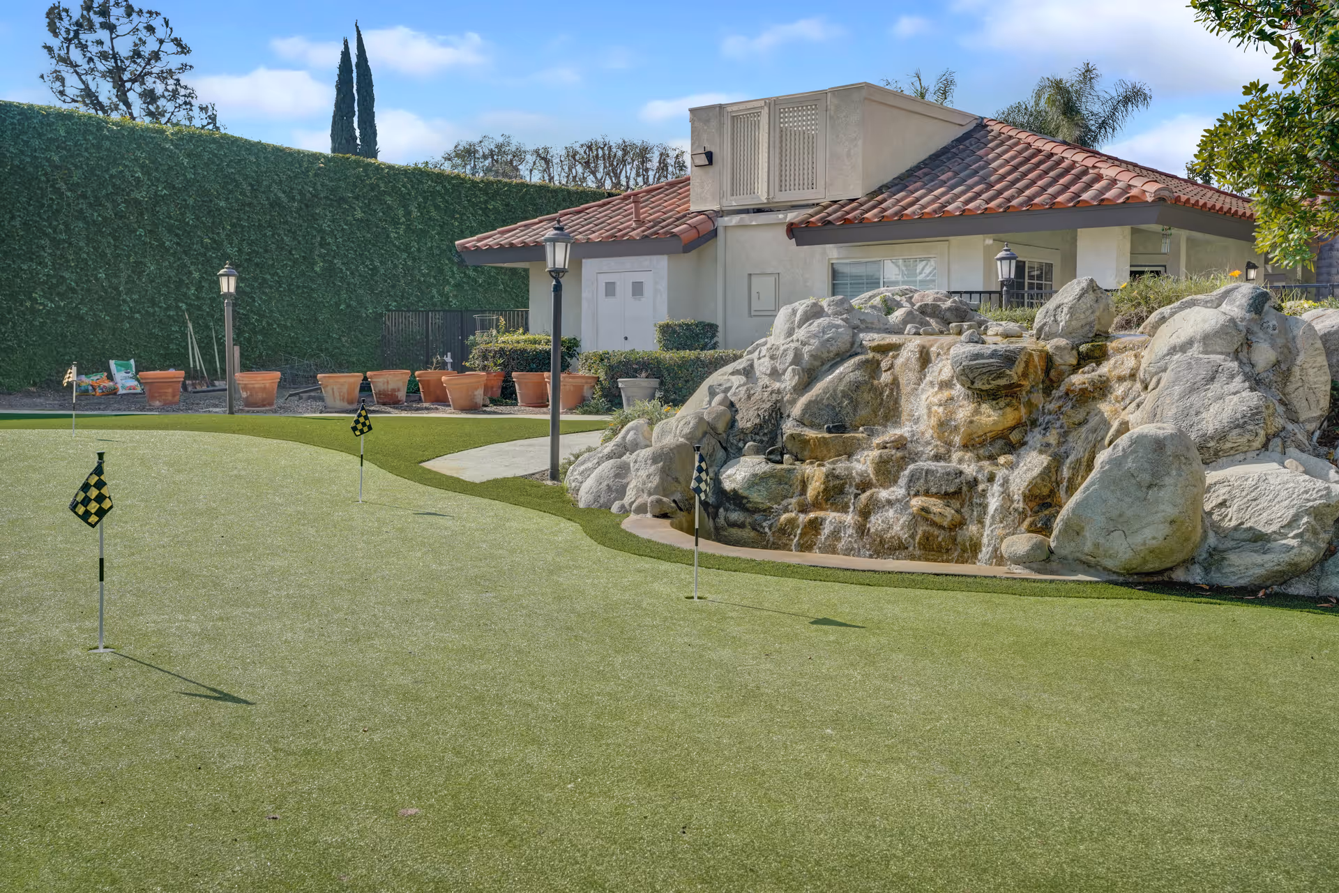 Outdoor putting green with small checkered flags, a rock waterfall feature, and a single-story building with a red tile roof.