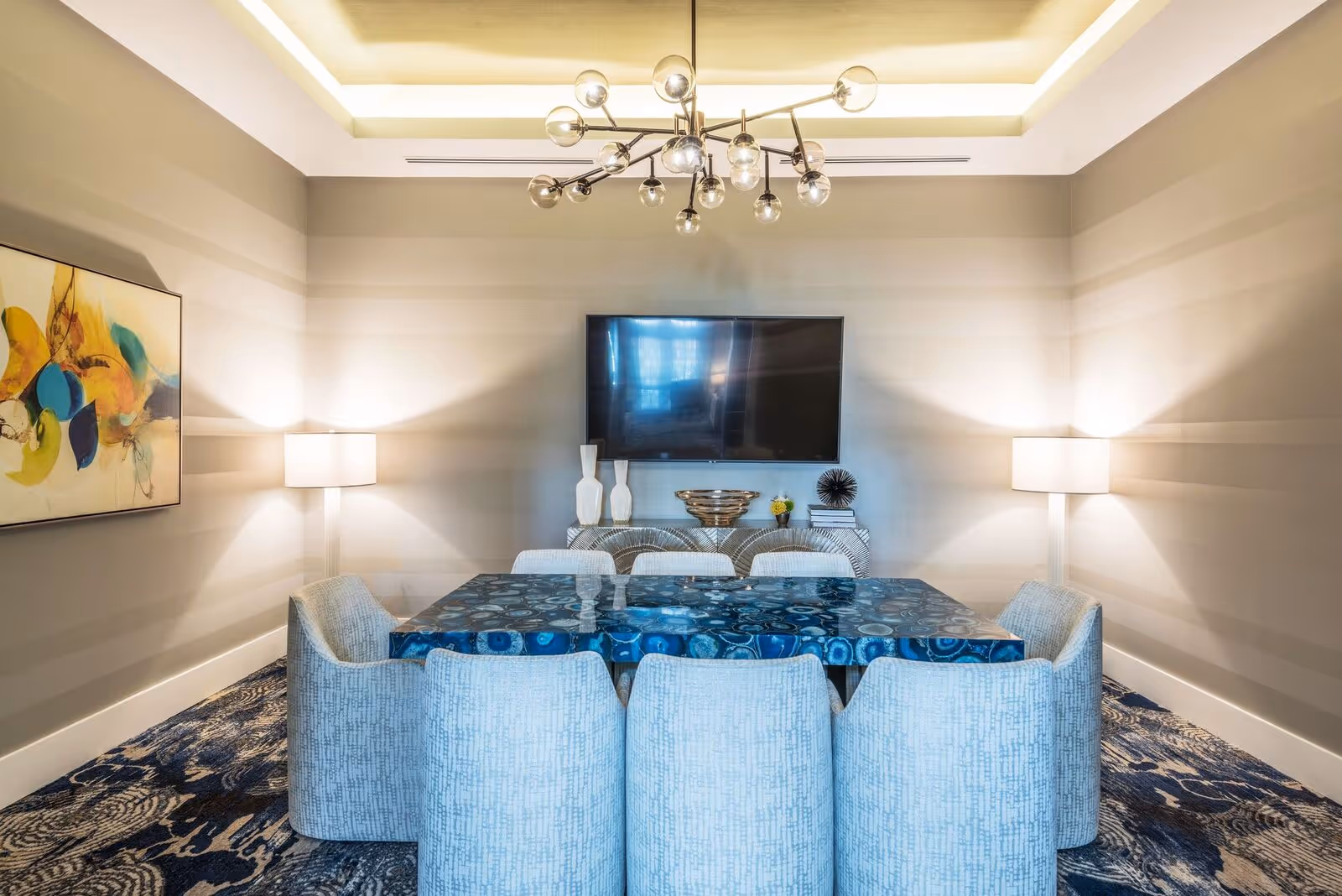 A modern dining room with a blue patterned rectangular table surrounded by eight light blue upholstered chairs. The room features striped beige walls, a large flat-screen TV mounted on the wall, two floor lamps on either side of the TV, a decorative sideboard with vases and a bowl, an abstract colorful painting on the left wall, and a contemporary chandelier with multiple exposed bulbs hanging from the ceiling.