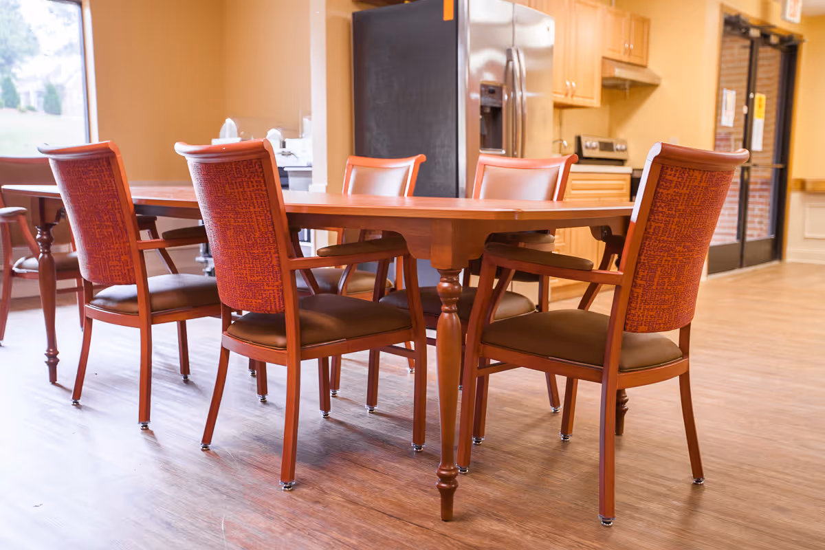 A dining area with a wooden table surrounded by six chairs with red patterned backs and brown seats. In the background, there is a kitchen area with wooden cabinets, a stainless steel refrigerator, and a stove. The floor is wooden, and there is a large window letting in natural light.