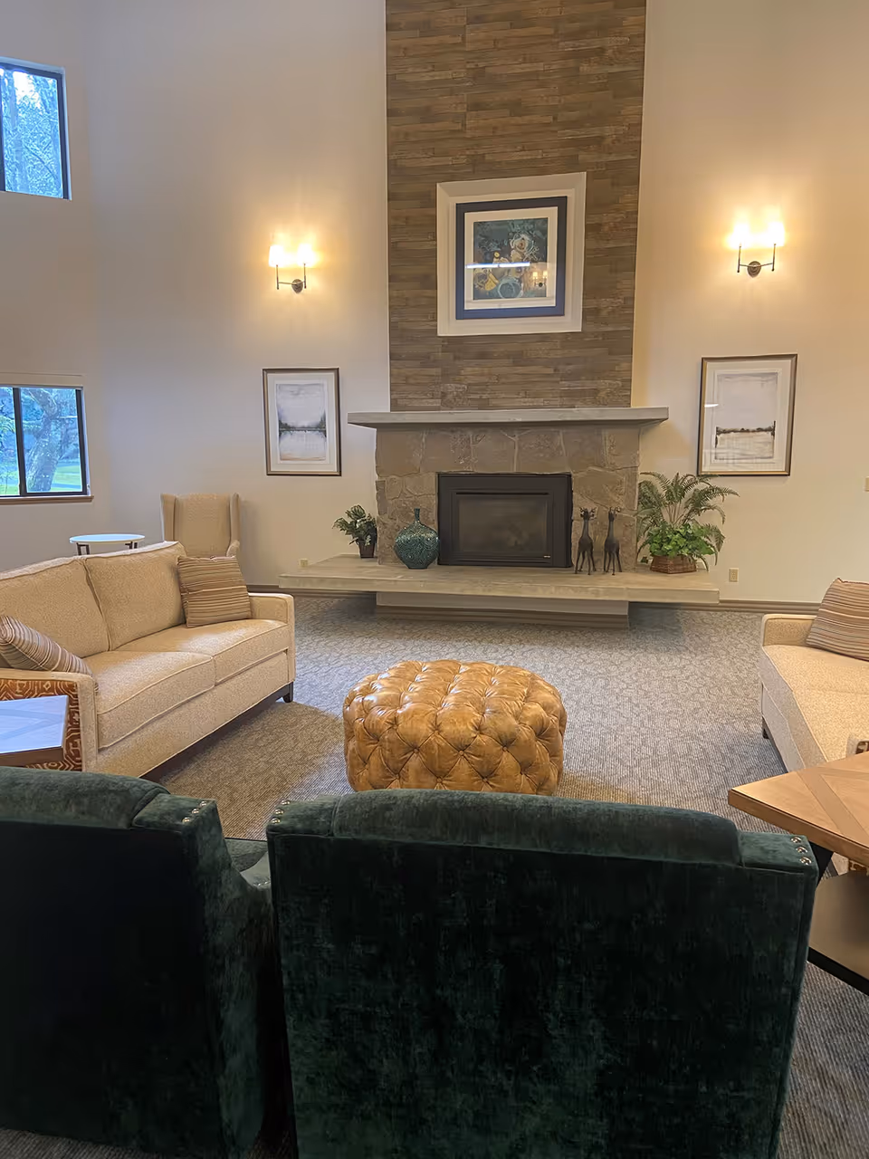 A cozy senior living room with a stone fireplace as the focal point. The room features two beige sofas with striped pillows, two dark green armchairs, a tan tufted ottoman in the center, and a beige wingback chair near a window. The fireplace mantel is decorated with plants, a blue vase, and two small giraffe figurines. Two framed pictures hang on either side of the fireplace, and two wall sconces provide warm lighting. Large windows let in natural light, and the room has a soft carpeted floor.