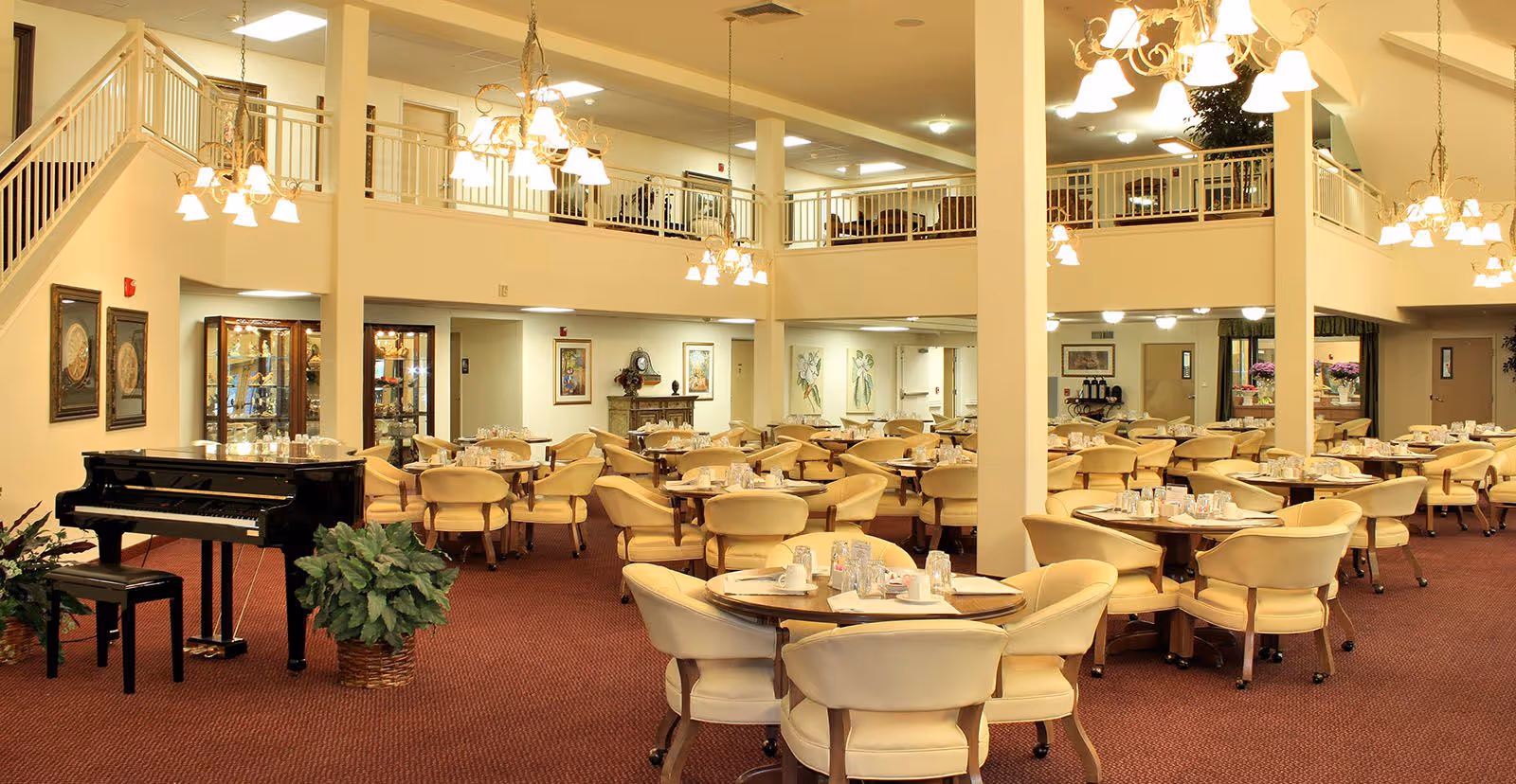 Spacious dining room with multiple round tables surrounded by cream-colored chairs. The room features a red carpet, chandeliers hanging from the ceiling, a black grand piano with a bench, and a second-floor balcony overlooking the area. The walls are decorated with framed artwork and plants are placed around the room.
