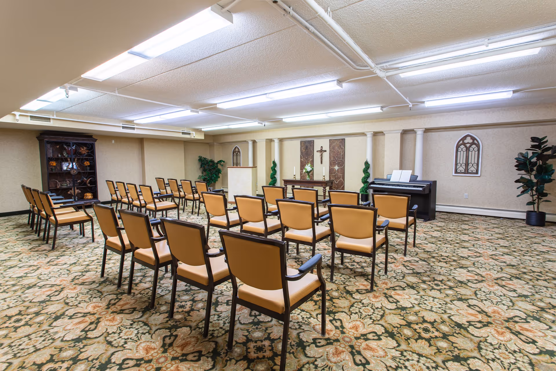 A small chapel or meditation room with rows of beige cushioned chairs arranged facing a wooden altar with a cross on the wall behind it. There is a piano with sheet music on the right side and decorative plants and a cabinet on the left. The room has patterned carpet and fluorescent ceiling lights.