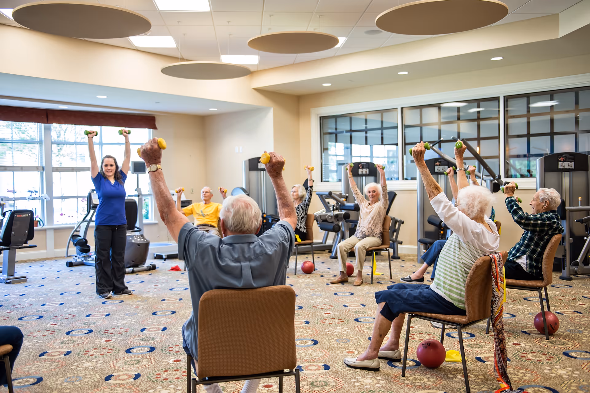 A group of elderly individuals seated in chairs in a fitness room, participating in a guided exercise class led by a female instructor. They are all holding small dumbbells and raising their arms above their heads. The room has large windows, exercise equipment, and a patterned carpet.