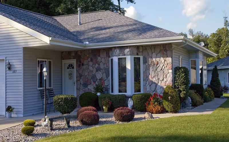Single-story building front with a stone and siding facade, a bay window, manicured shrubs and a lawn.