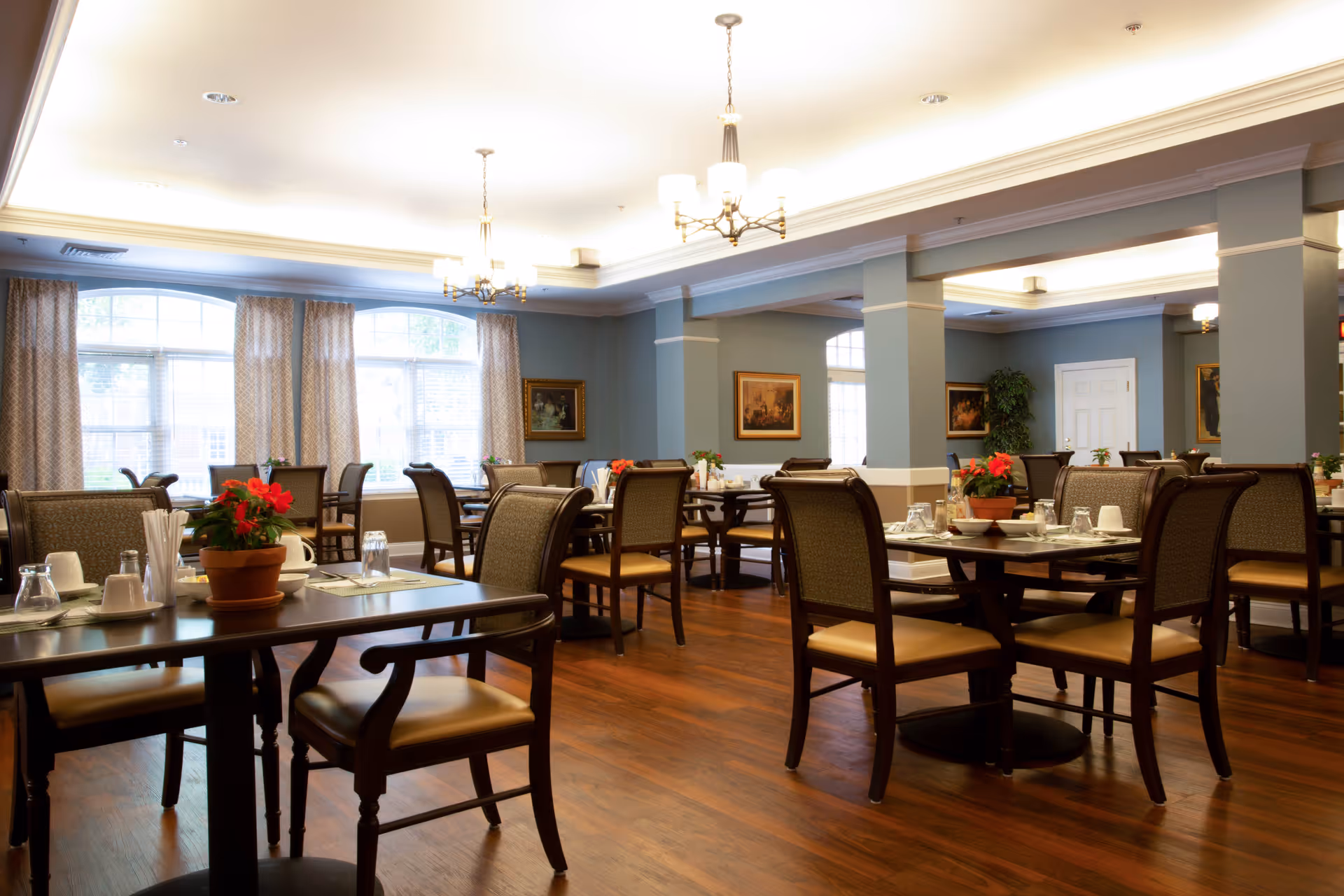 A spacious dining room with multiple wooden tables and chairs arranged neatly. Each table is set with glasses, cups, and napkins, and some have potted red flowers as centerpieces. The room has large windows with curtains allowing natural light to brighten the space. The walls are painted light blue with white trim, and there are framed paintings hanging on the walls. Ceiling lights with chandeliers provide additional lighting.