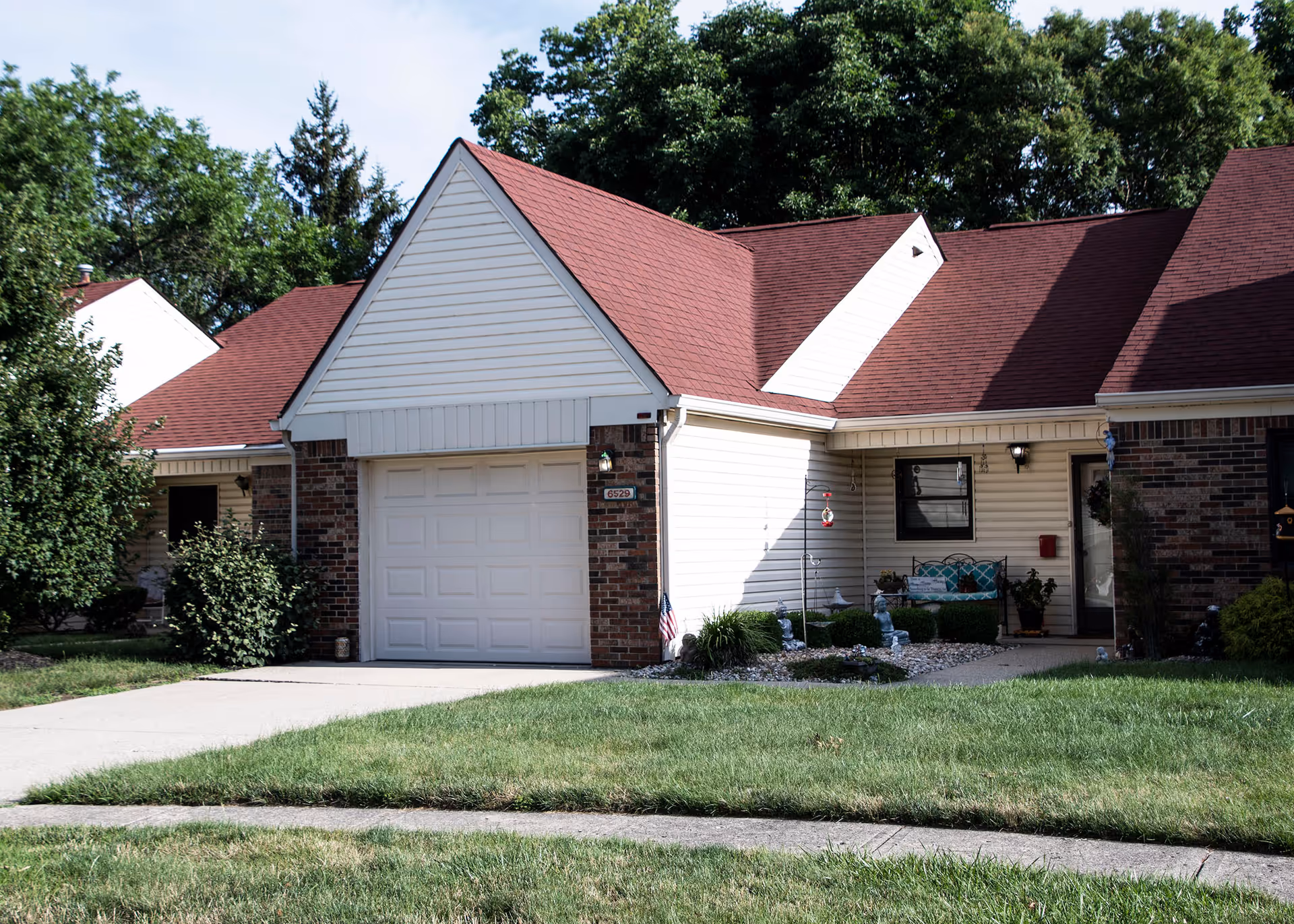 Exterior view of a single-story senior living unit with a red roof, white siding, and brick accents. The unit features a garage door, a small front porch with a bench, potted plants, and decorative garden statues. There is a concrete driveway and a well-maintained lawn in front of the unit, with trees and shrubs in the background.