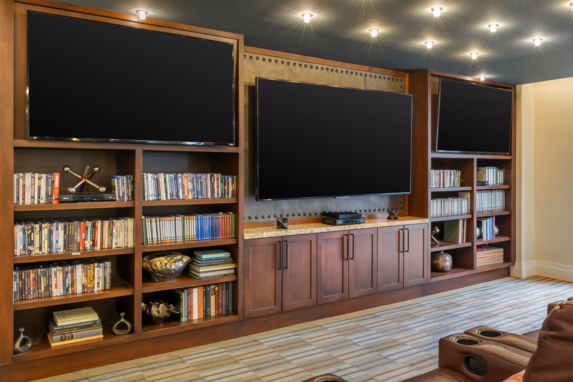Cozy media room with built-in wooden shelving filled with books and DVDs, three large flat-screen TVs mounted above cabinets, and recliner seating.