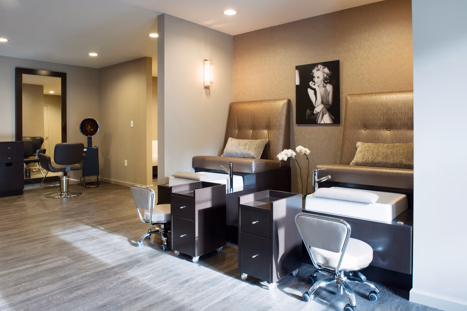 Well-lit salon area with two pedicure chairs, manicure stations, rolling stools, and a framed black-and-white portrait on the wall.