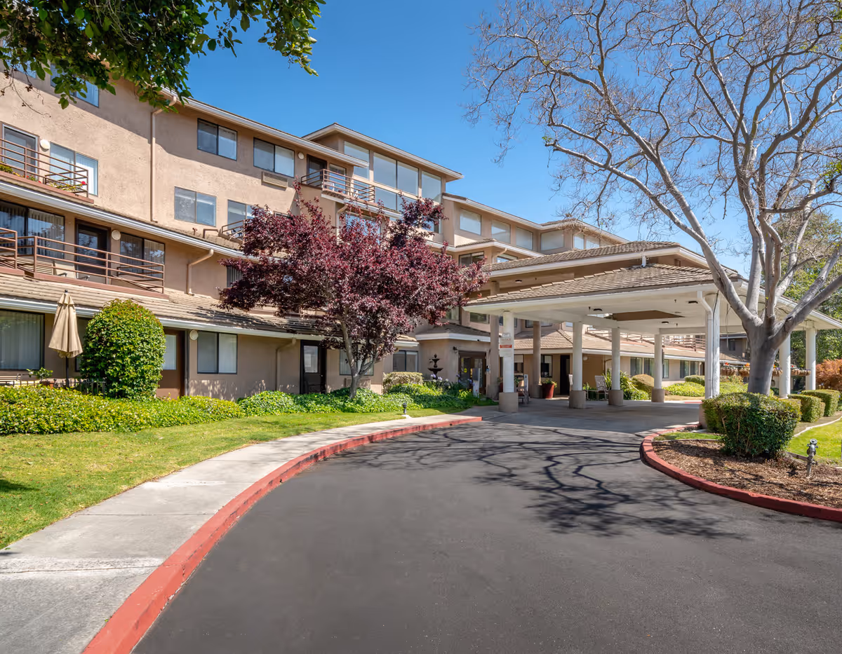 Exterior view of a multi-story retirement community building with balconies, a covered entrance driveway, landscaped greenery, and trees under a clear blue sky.