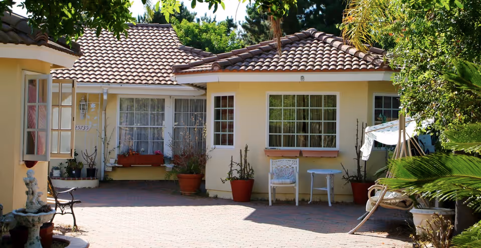 Exterior view of a single-story yellow building with a tiled roof, several windows, potted plants, a white chair with a cushion, a small white table, and a swing covered with a canopy. The area is paved with bricks and surrounded by greenery.