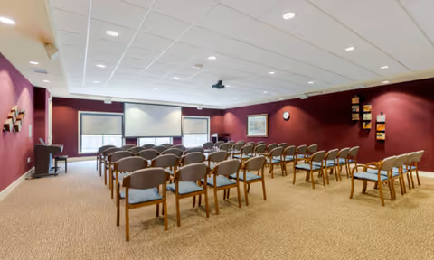 Conference room with rows of wooden chairs facing two projection screens against maroon walls.